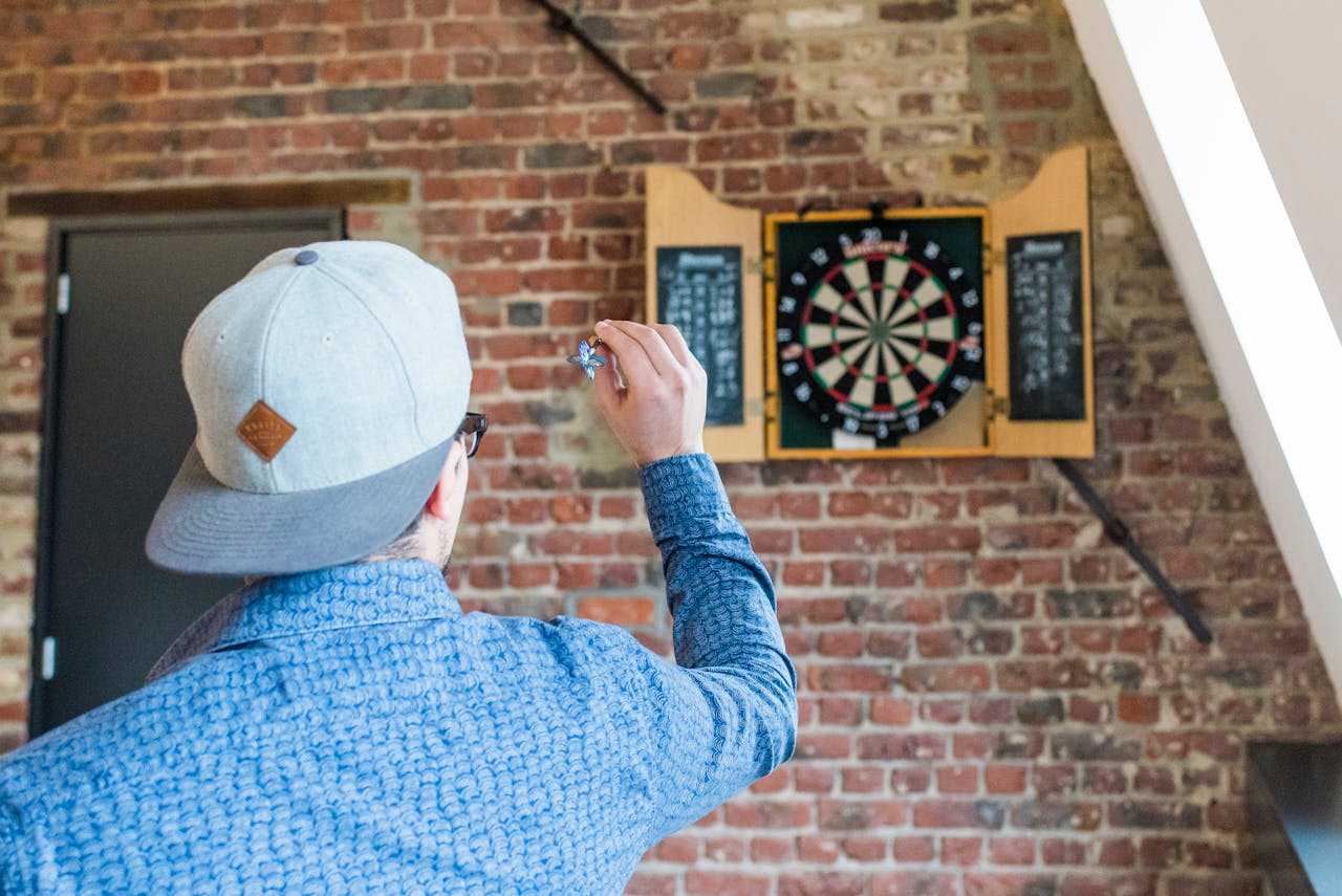 why-choose-us An adult man in a blue shirt playing darts indoors, aiming at a brick wall-mounted dartboard.